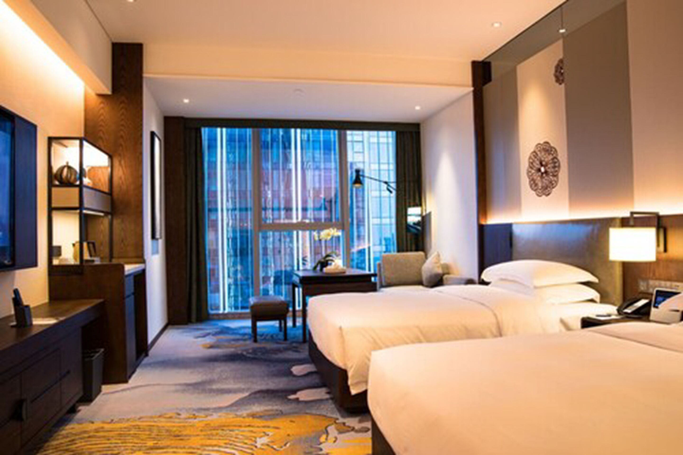 A hotel room with two beds, a desk, a chair, a large window with curtains, and a decorative wall piece.