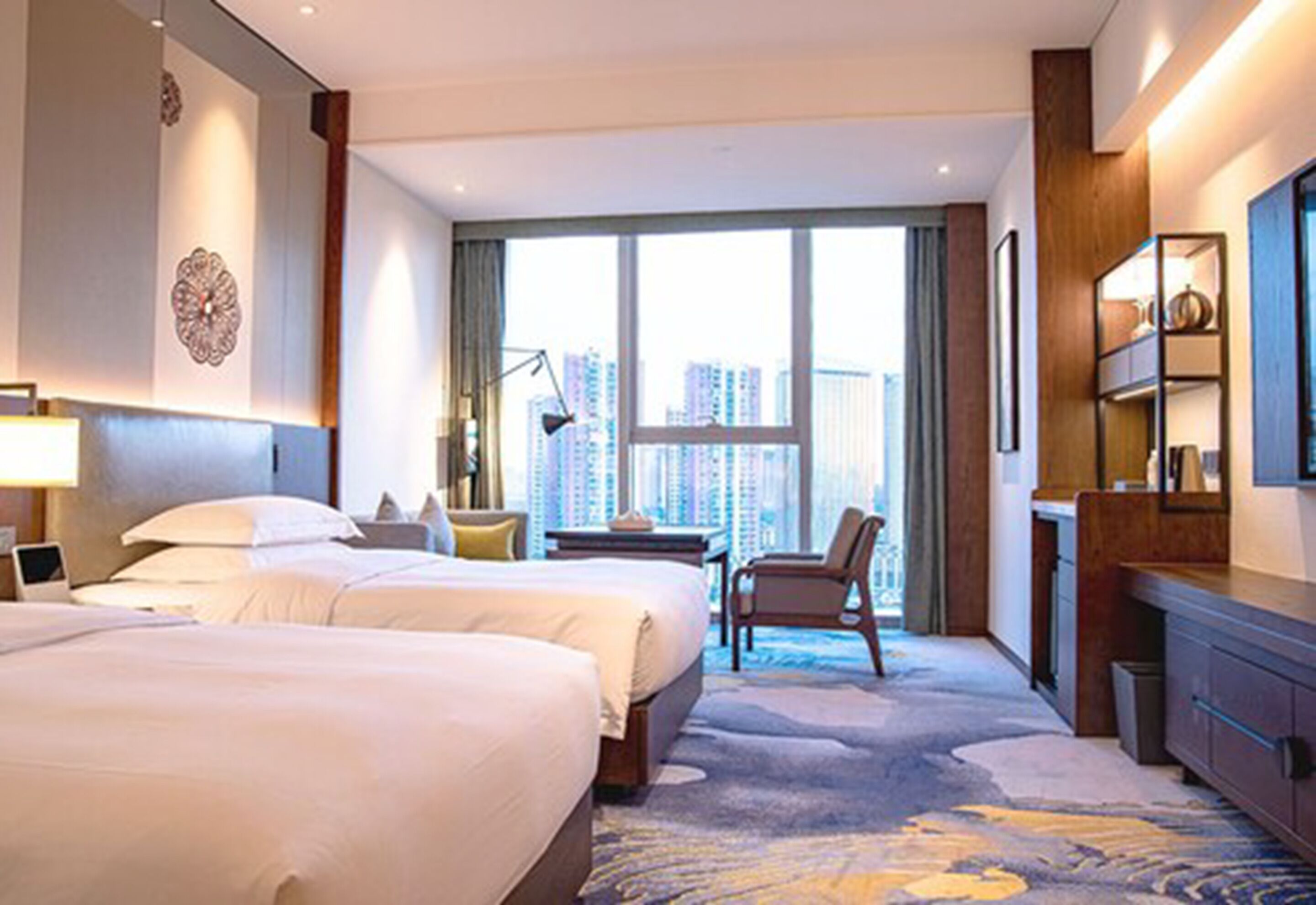 A hotel room with two beds, a desk, a chair, a large window with a city view, and a bookshelf.