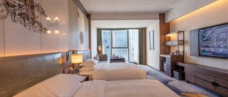 A hotel room with two beds, a desk, a chair, a TV, and a large window with a view of buildings.