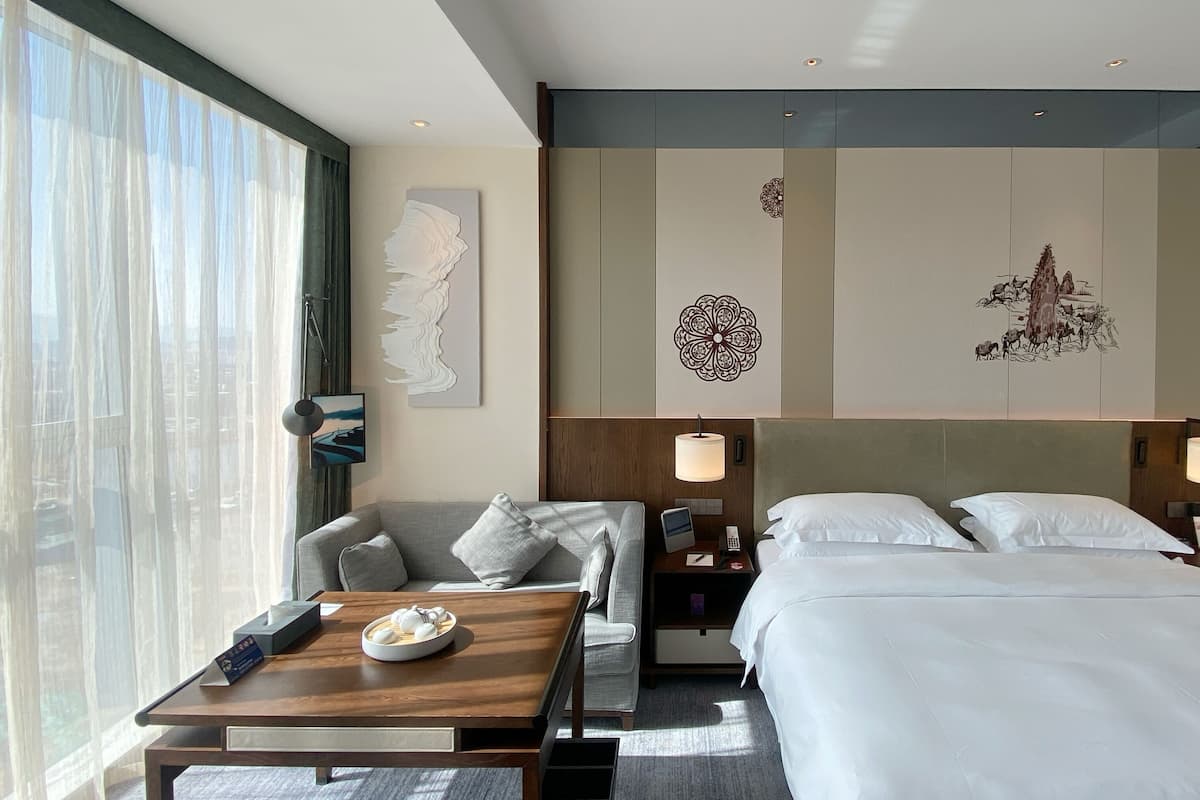 A hotel room with a large bed, a sofa, a desk with a bowl of fruit, and artwork on the walls.