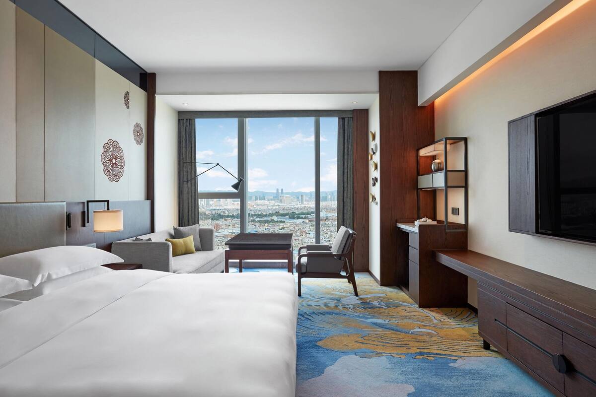 A hotel room with a large bed, a desk, a chair, a TV, and a view of the city.