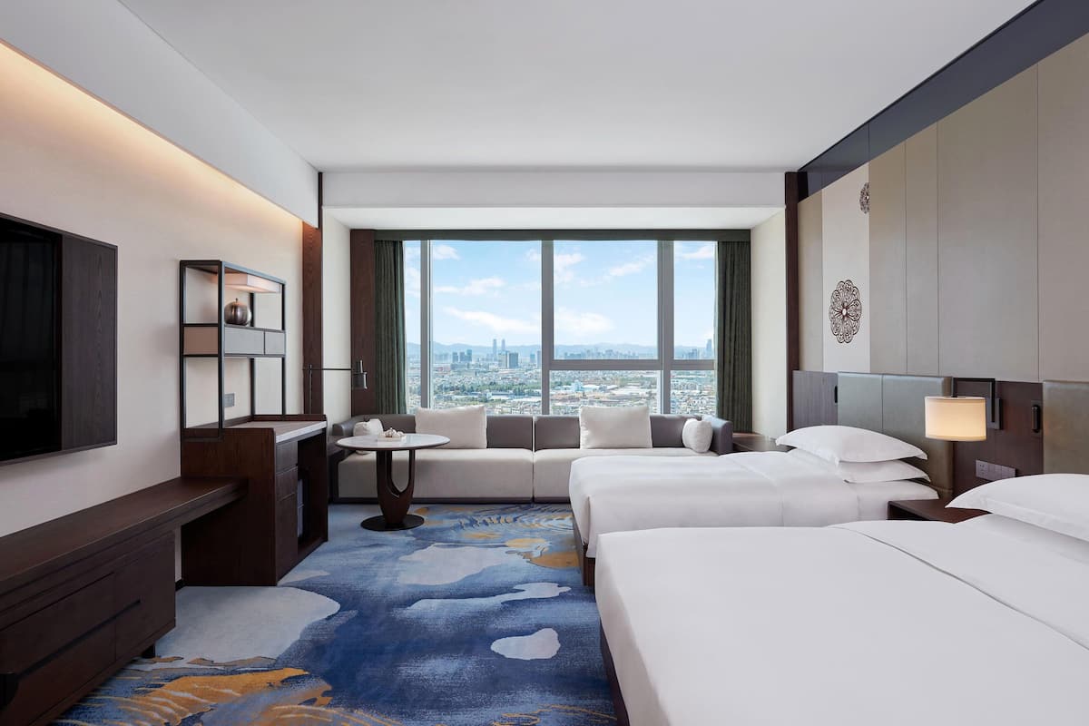A hotel room with a city view, a sofa, two beds, a desk, and a TV.