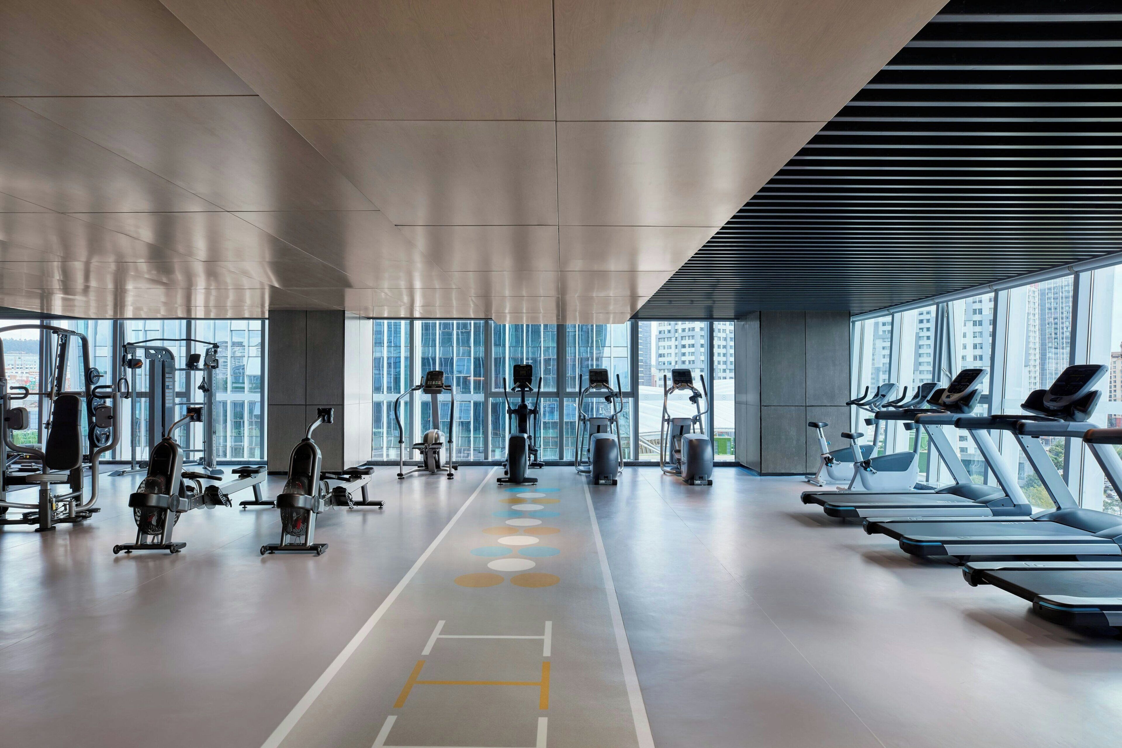 A modern gym with treadmills and weight machines, large windows, and a high ceiling.