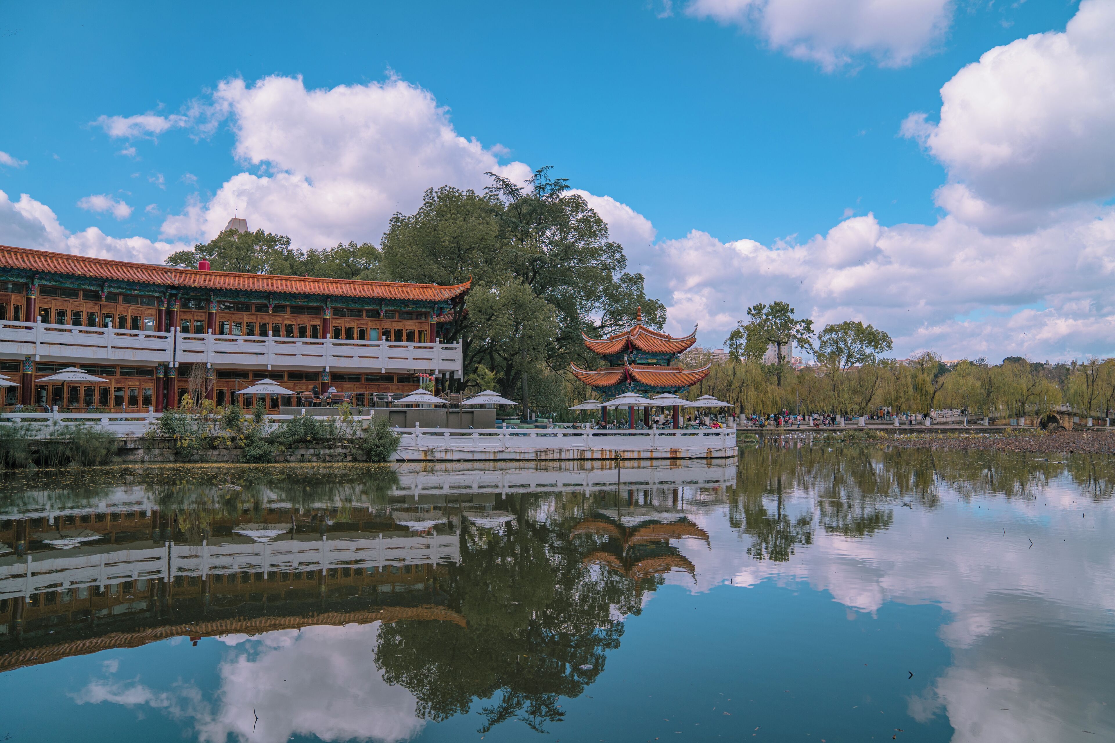 A traditional building with a reflection in a calm body of water, surrounded by trees and a clear sky.