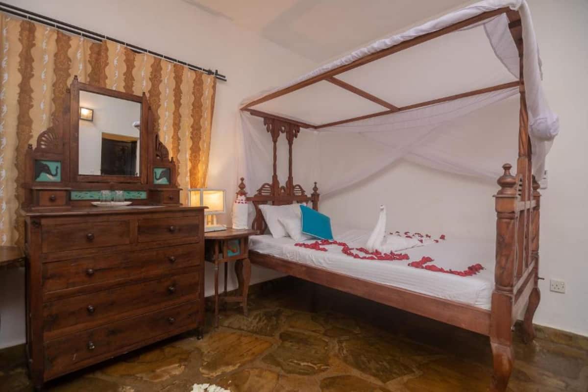 Una cama con dosel de madera y cuatro columnas, un aparador de madera con cajones y un espejo con detalles decorativos.