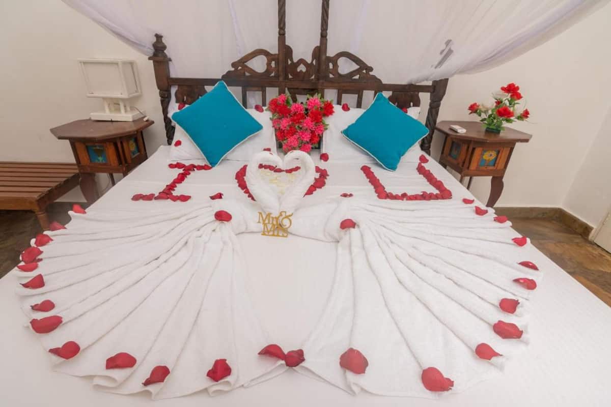 Una cama con pétalos de rosa formando un corazón y un diseño de cisne, flanqueada por dos almohadas azules y un ramo de flores rojas y blancas.