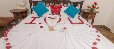 Una cama con pétalos de rosa formando un corazón y un diseño de cisne, flanqueada por dos almohadas azules y un ramo de flores rojas y blancas.