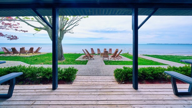Ein überdachter Außenbereich mit Holzbänken und Tischen, ein Betonweg zum Strand und Blick aufs Meer.