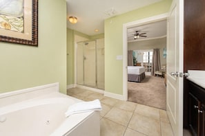 A bathroom with a bathtub, a shower, and a glass door leading to a bedroom with a bed and a chair.