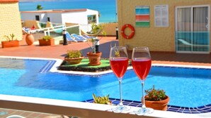Un espace aménagé au bord d’une piscine, avec deux verres contenant un liquide rouge, des plantes en pot et une bouée de sauvetage accrochée à un mur du bâtiment.