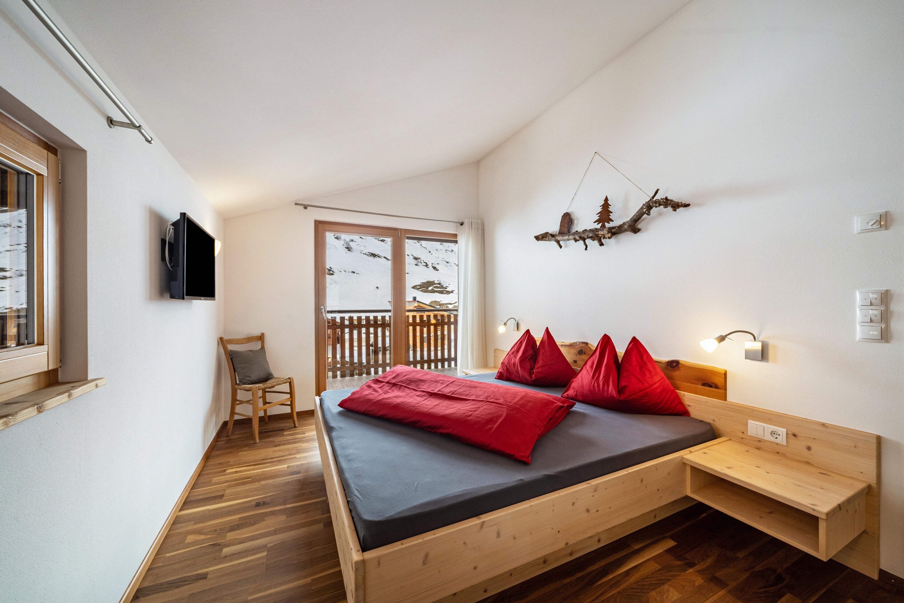 Ein Schlafzimmer mit Bett, Sessel, Fenster mit Schneeausblick, Wand-Fernseher und einer hölzernen Bank.