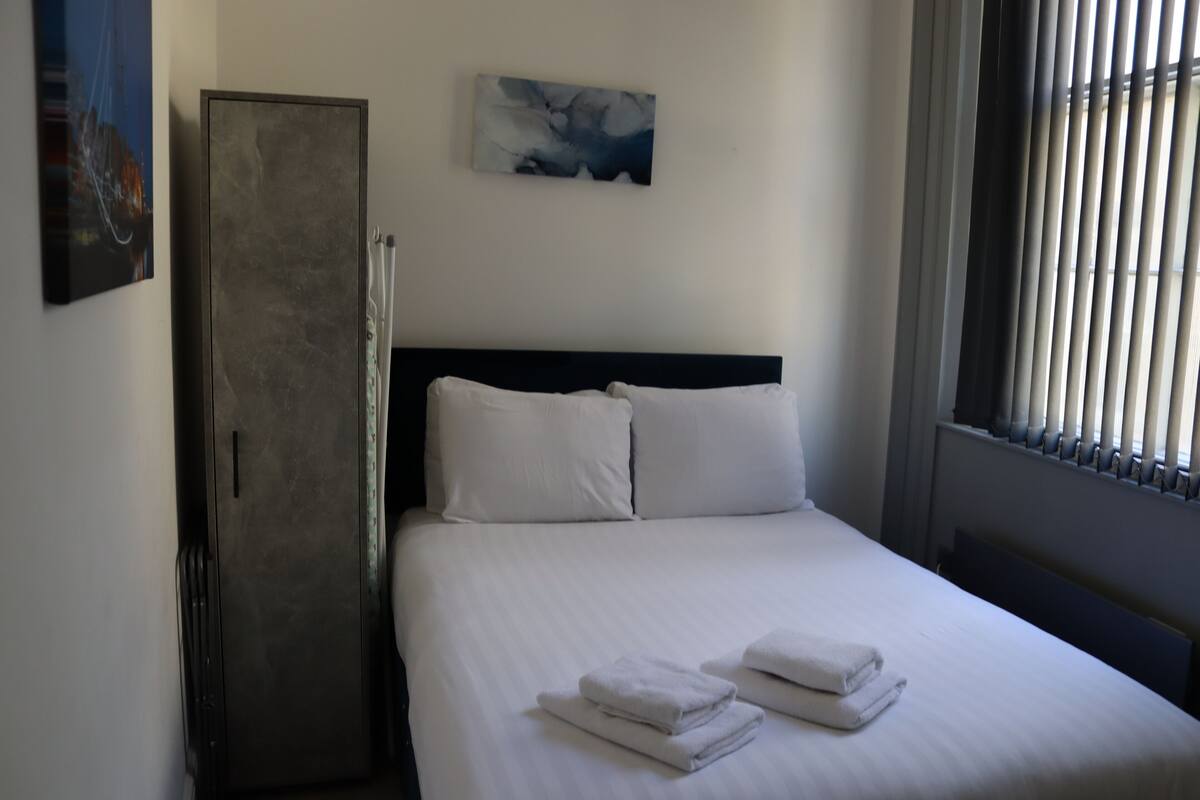 A neatly made bed with white linens and pillows, a dark headboard, and a painting on the wall.