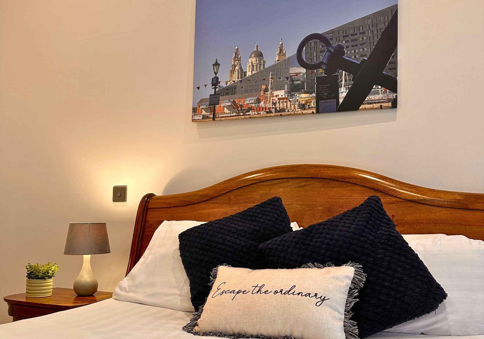 Ein Hotelzimmer mit einem hölzernen Kopfteil, einem Bett mit weißen und dunkelblauen Kissen, einer Lampe und einem an der Wand befestigten Stadtbild.