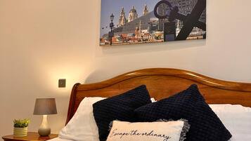 Ein Hotelzimmer mit einem hölzernen Kopfteil, einem Bett mit weißen und dunkelblauen Kissen, einer Lampe und einem an der Wand befestigten Stadtbild.