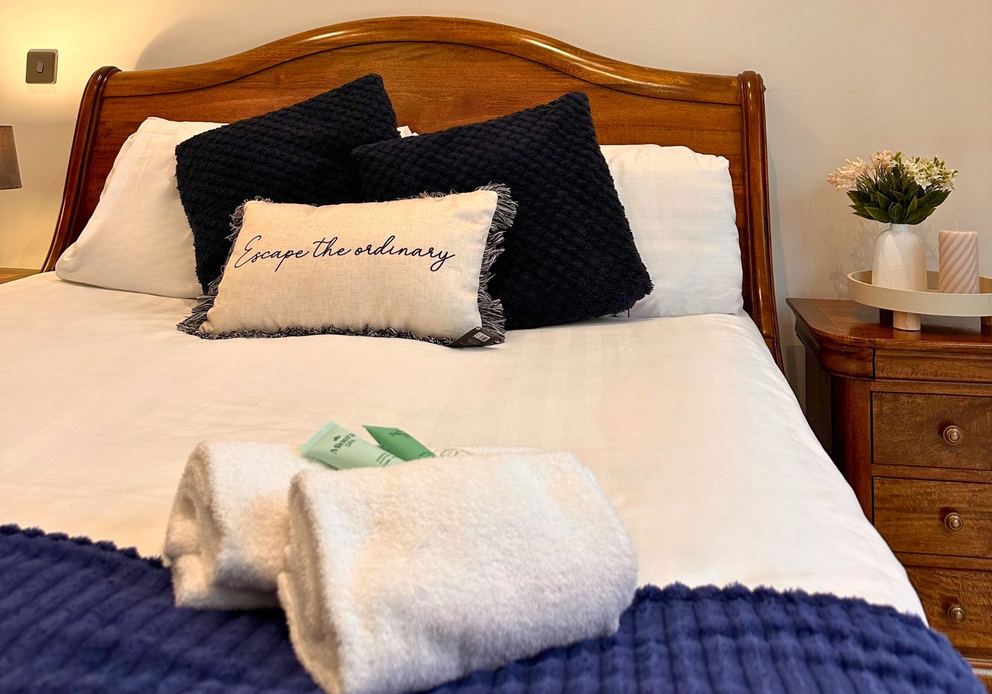 Una cama bien tendida con una almohada que dice “Escapa de lo común”, una toalla doblada y un pequeño florero con flores sobre una mesita de noche.