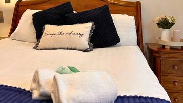 Una cama bien tendida con una almohada que dice “Escapa de lo común”, una toalla doblada y un pequeño florero con flores sobre una mesita de noche.