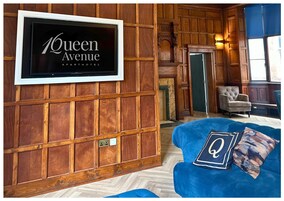 A hotel room with a large TV mounted on a wooden paneled wall, a blue sofa with a decorative pillow, and a classic armchair.