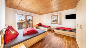 A room with wooden flooring, a bed with red bedding, a small table, a TV mounted on the wall, and large windows with a view of a building outside. - Apartment "Enzian" on a Mountain Farm with Balcony (Moos in Passeier)