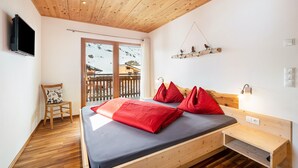 A bedroom with a wooden ceiling, a bed with red pillows, a wooden nightstand, a TV mounted on the wall, and a balcony with a view of a snowy landscape. - Apartment "Enzian" on a Mountain Farm with Balcony (Moos in Passeier)