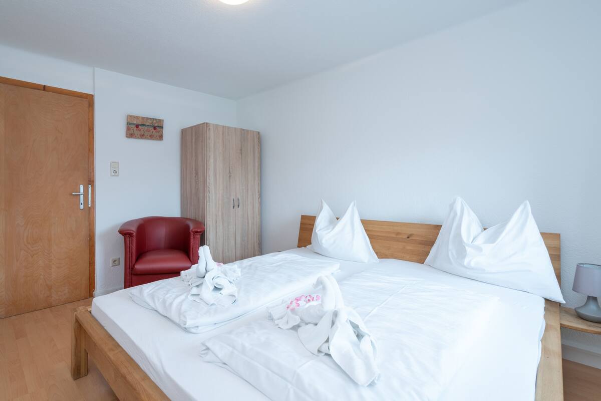 A hotel room with a wooden bed, white linens, a red chair, and a wooden wardrobe.