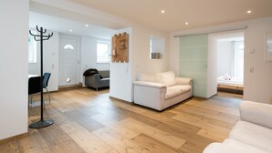 Een ruime woonkamer met houten vloer, een witte bank, een grijze fauteuil en een kapstok.
