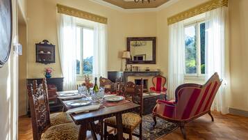 Uma sala de jantar com uma mesa de madeira posta para quatro pessoas, rodeada por cadeiras ornamentadas, uma lareira e um espelho grande na parede.