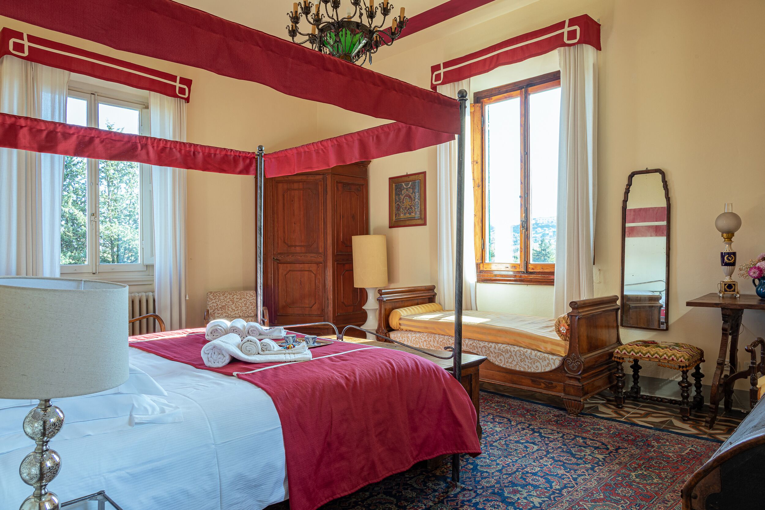 Uma cama de dossel com dossel vermelho, uma mesinha de cabeceira de madeira, uma mesa de apoio com abajur e uma janela grande com cortinas.