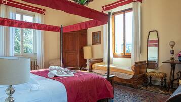Uma cama de dossel com dossel vermelho, uma mesinha de cabeceira de madeira, uma mesa de apoio com abajur e uma janela grande com cortinas.