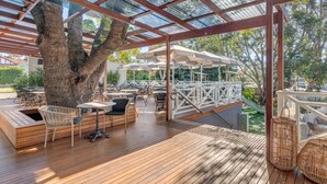An outdoor seating area with wooden tables and chairs, a large tree, and a covered patio with umbrellas. - Wentworth Hotel (Homebush)