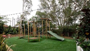 A playground with a slide and climbing structures on artificial grass, surrounded by trees and plants. - Wentworth Hotel (Homebush)