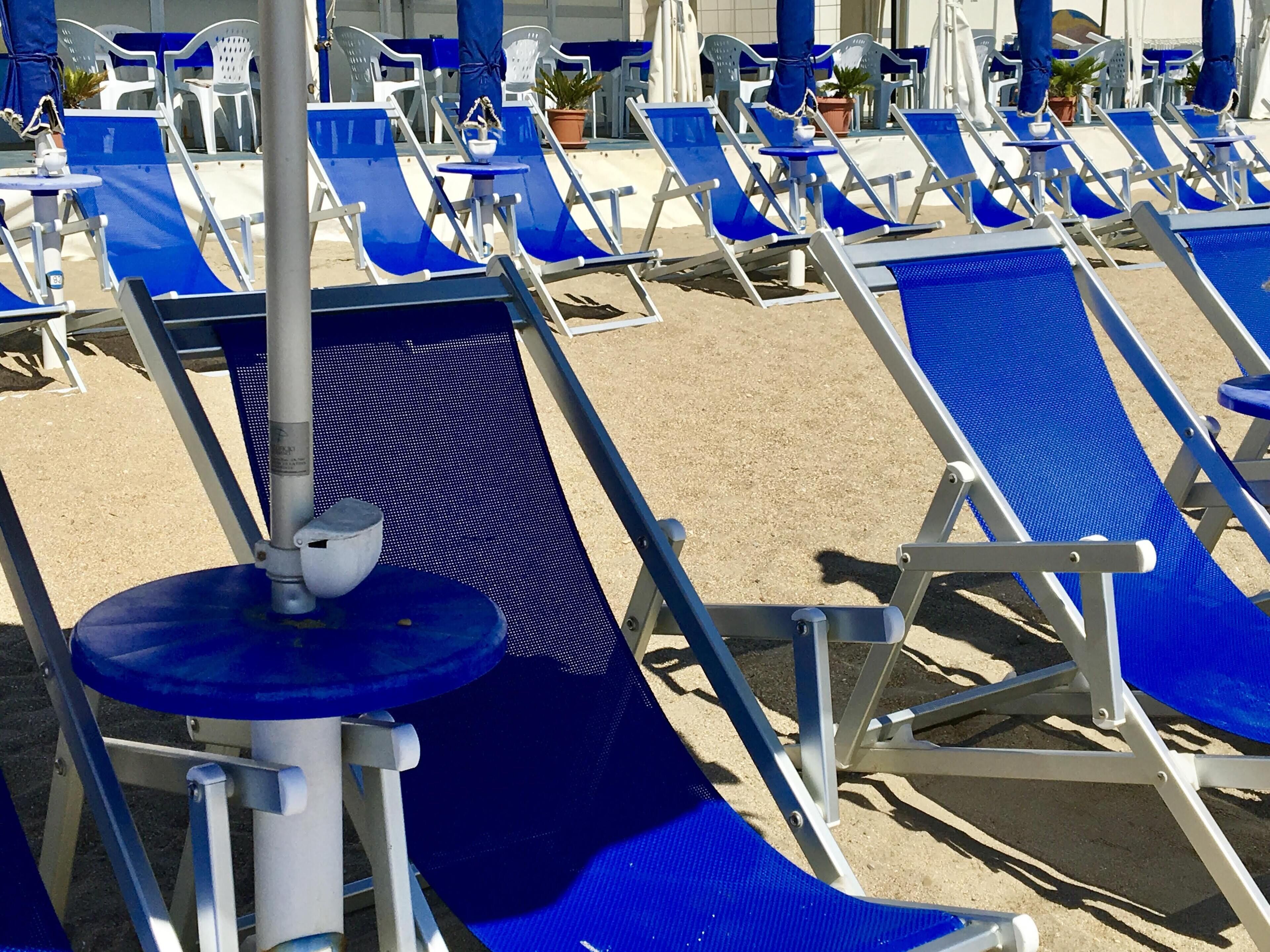 De longues rangées de chaises longues et de transats bleus sur une plage de sable, agrémentées de quelques plantes en pot et de parasols.