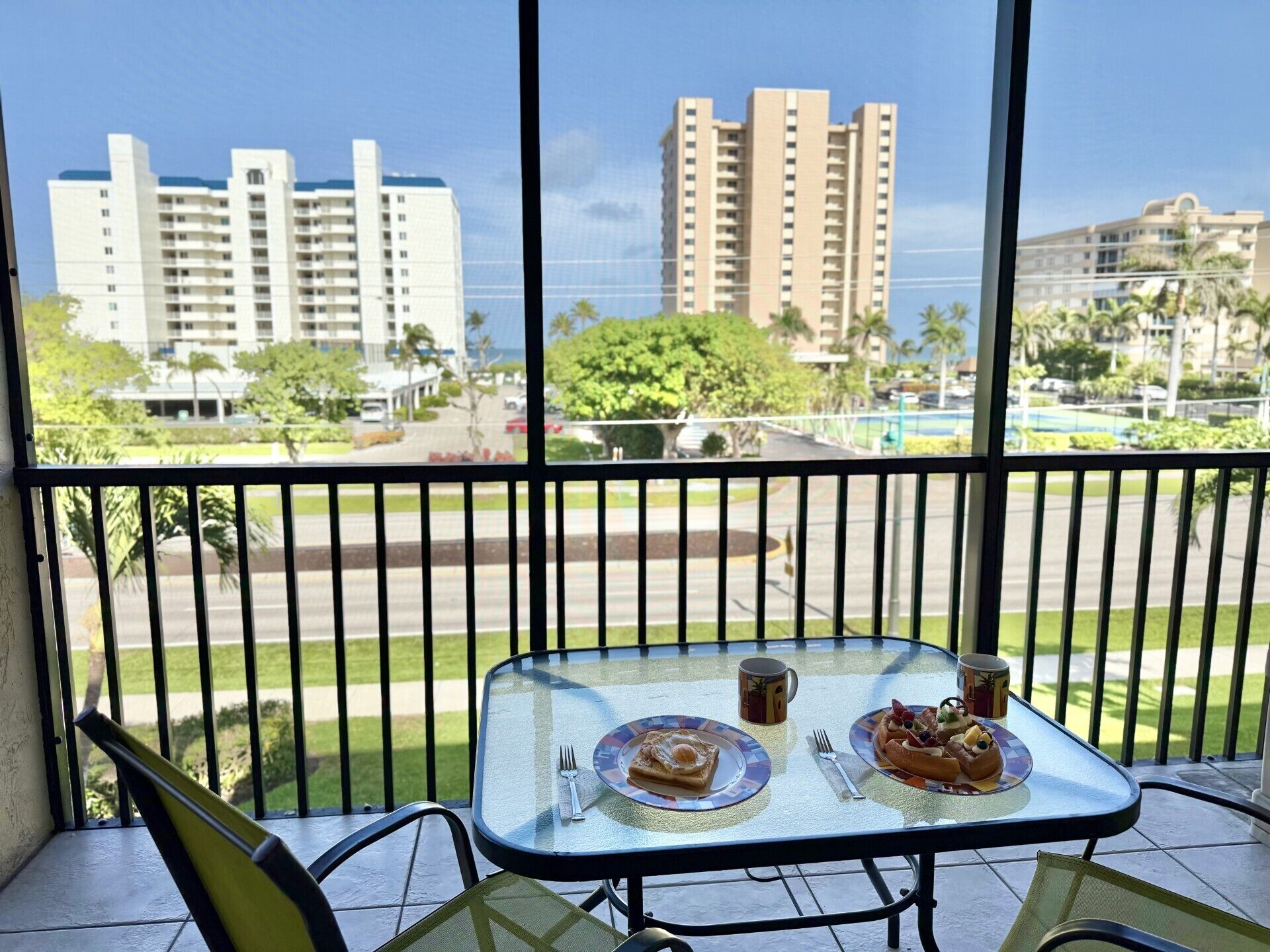 A balcony with a table set for breakfast, overlooking a cityscape with tall buildings and a clear sky.