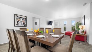 A dining area with a wooden table set for four, surrounded by chairs. There is a television mounted on the wall, a red sofa, and a small table with a lamp and plant. - ModernPrivatePoolQuiet3BR2BA Eagle/Palm Beach (Noord)