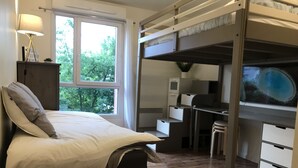 Une chambre avec un lit superposé, un bureau, une lampe et une vue sur les arbres par la fenêtre.
