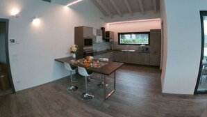 A modern kitchen with a dining area, featuring a central island, bar stools, and a large window providing natural light. - Villa Vittoria - POOL  (Colico Piano)