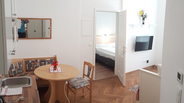 Un ambiente compatto con angolo cottura, zona pranzo con tavolo e sedie in legno, un letto con testiera e una TV a parete.
