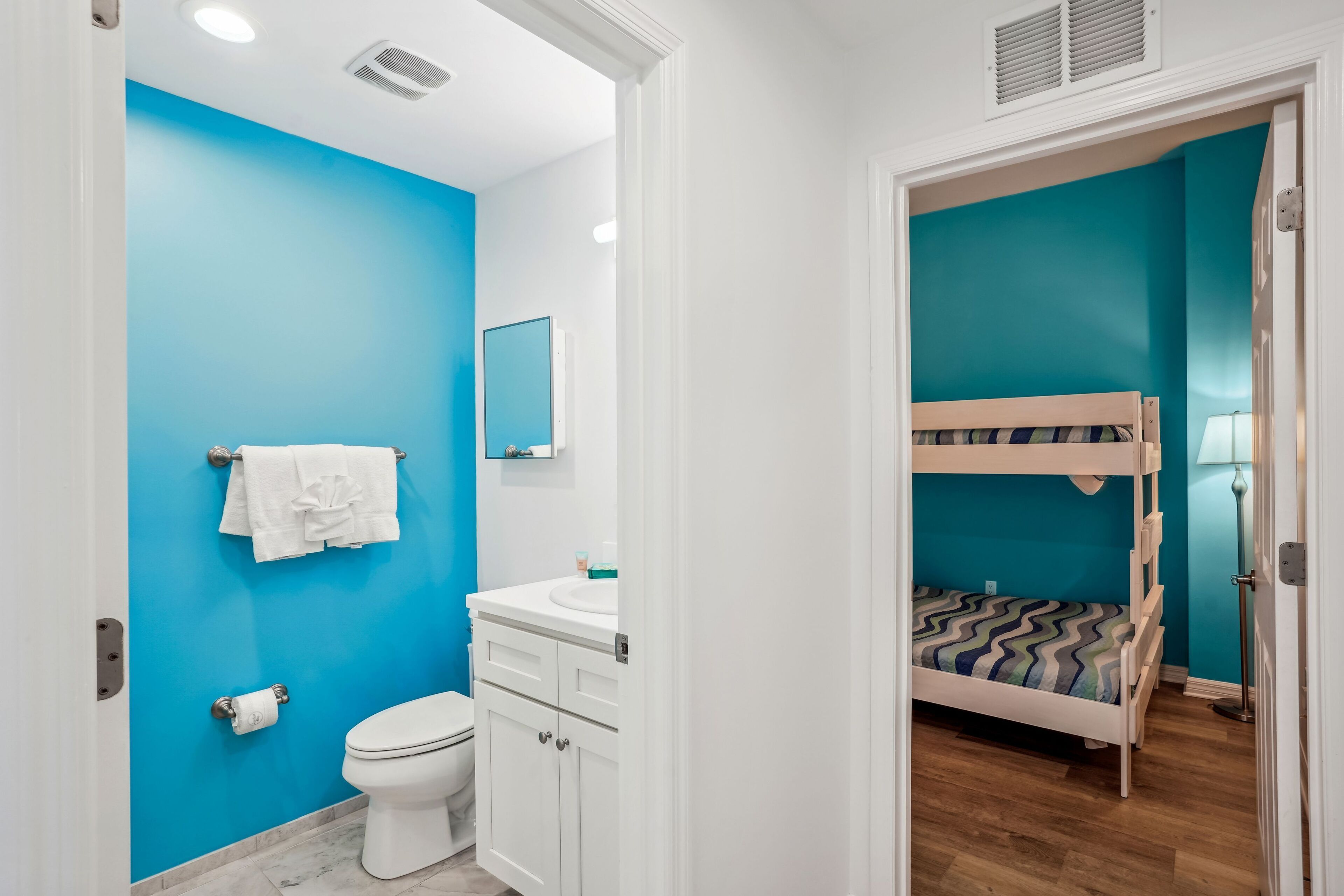 Une salle de bain avec un mur bleu, un lavabo blanc et des toilettes. La pièce adjacente contient un lit superposé.