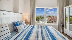 A bedroom with a large bed, a nightstand, a lamp, and a view of a marina through a window.