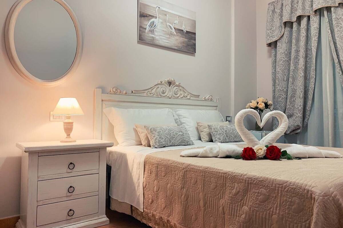 A bedroom with a bed, a bedside table with a lamp, a mirror, and a painting on the wall.