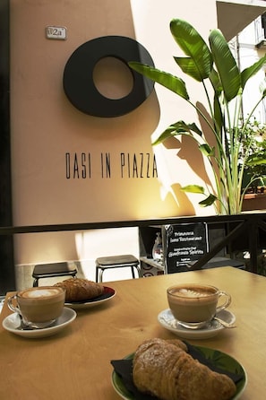 A table with two cups of coffee and two croissants, a sign reading "OASI IN PIAZZA", and a large plant. - B&B Vico d'Ercole (Fondi)