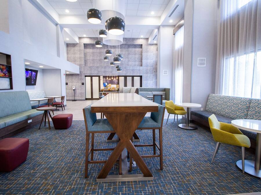 Eine moderne Hotellobby mit einem großen Holztisch, blau-gelben Stühlen, einem roten Sofa und einer Sitzecke mit einem gemusterten Teppich.