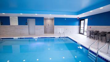 Una piscina con agua azul cristalina, rodeada de paredes azules y suelo de baldosas. Hay barandillas de metal a lo largo del borde de la piscina, y una puerta que conduce a los vestuarios.