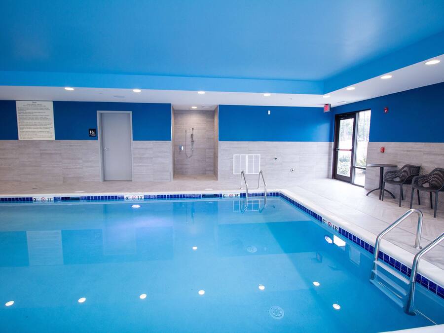 Una piscina con agua azul cristalina, rodeada de paredes azules y suelo de baldosas. Hay barandillas de metal a lo largo del borde de la piscina, y una puerta que conduce a los vestuarios.