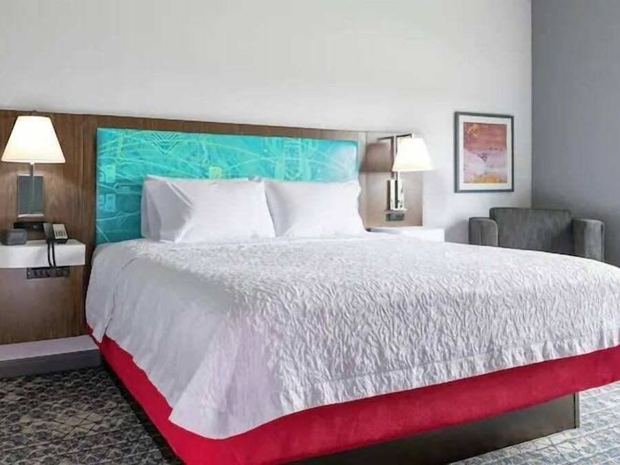 Habitación de hotel con una cama grande, dos mesitas de noche con lámparas, un cabecero con diseño abstracto azul, un sillón gris y un cuadro en la pared.