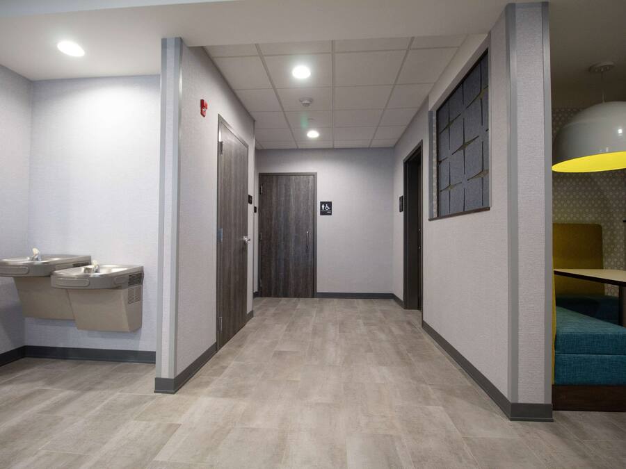 A hallway with a light wood floor, a wall-mounted sink, a door, and a seating area with a yellow pendant light.