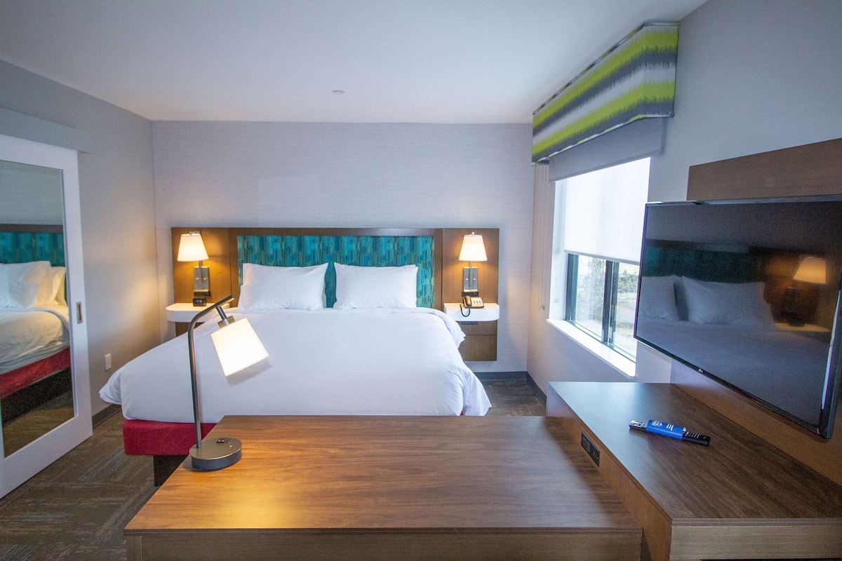 A hotel room with a bed, bedside lamps, a TV, a desk, and a window.