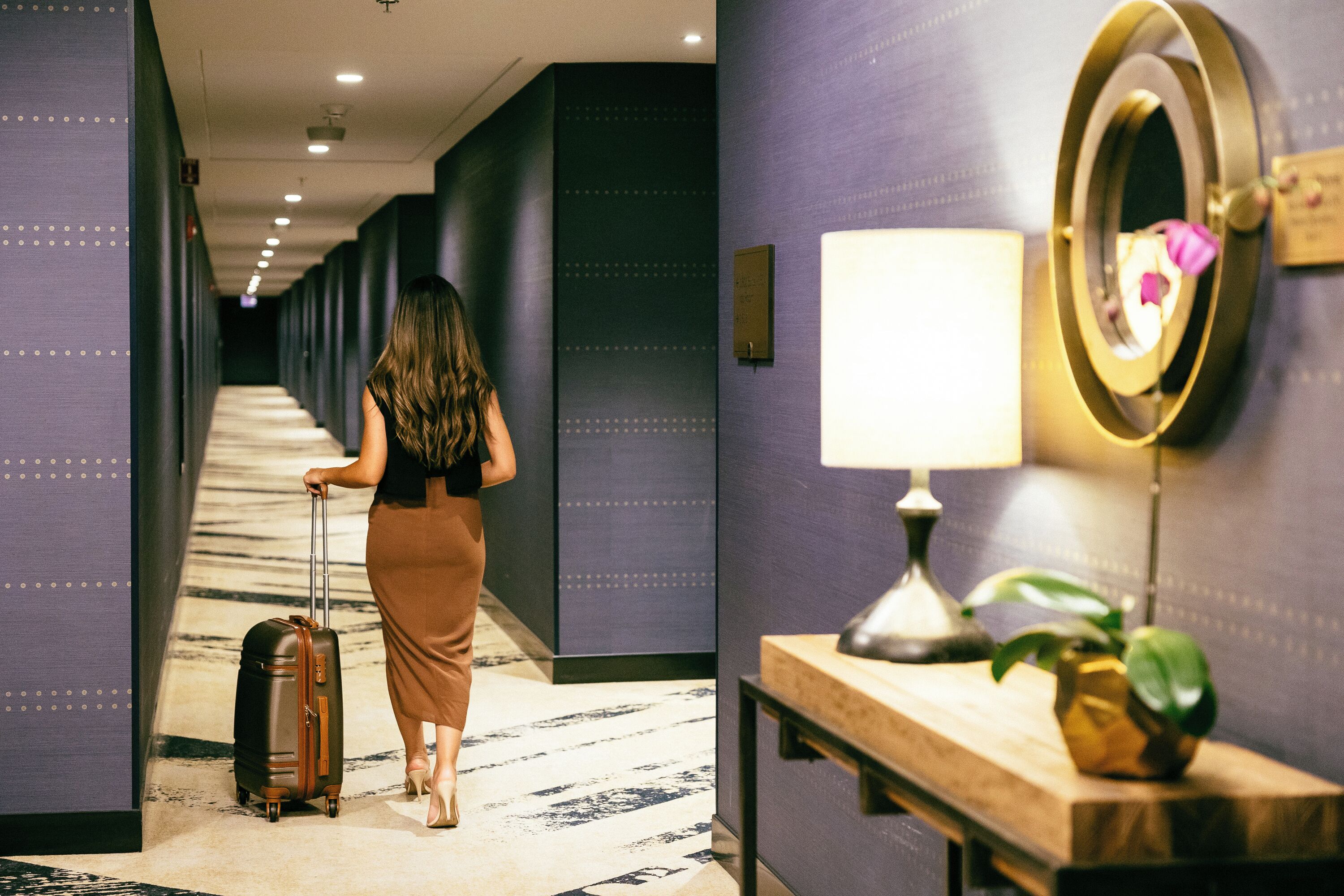 A hotel corridor with a woman pulling a suitcase, a wall-mounted mirror, and a bedside table with a lamp and a plant.