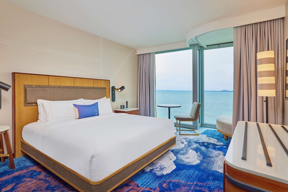 A hotel room with a large bed, a desk, a chair, a small table, and a view of the sea through a large window.
