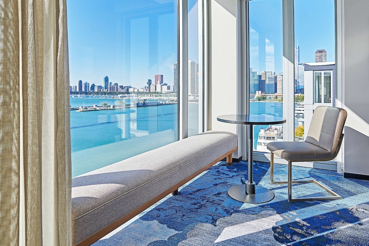 A hotel room with a large window, a round table, a chair, and a bench.