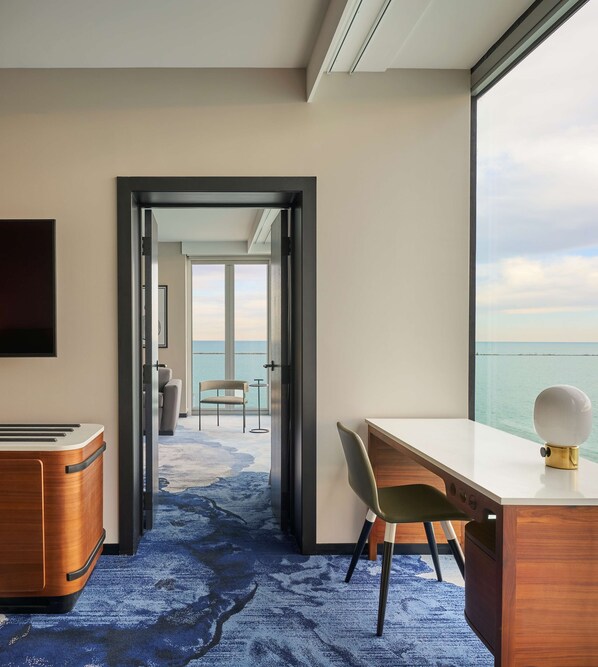 Une chambre d’hôtel dotée d’une grande fenêtre donnant sur la mer, d’un bureau avec une lampe, d’une chaise et d’une petite table.